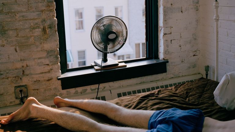 man in boxers lying face down on a bed with a fan blowing at him